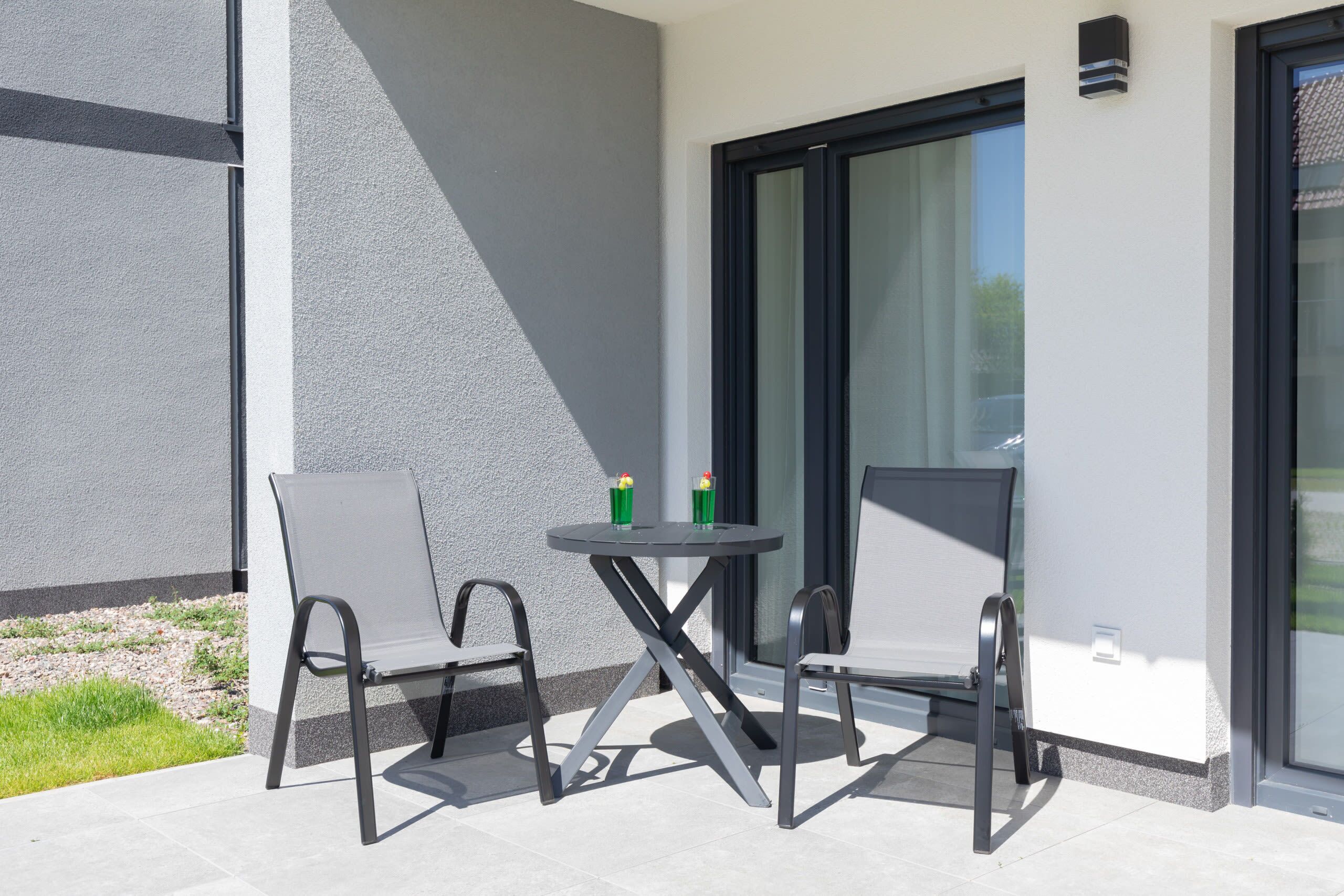 A patio with a table and two chairs, a small table with two cups, and a building with a glass door and window.