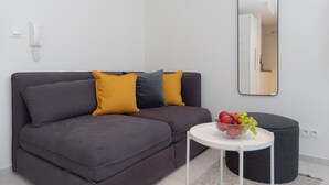 A modern living room with a grey sofa, a round black ottoman, a small white table with a fruit bowl, and a large mirror on the wall.