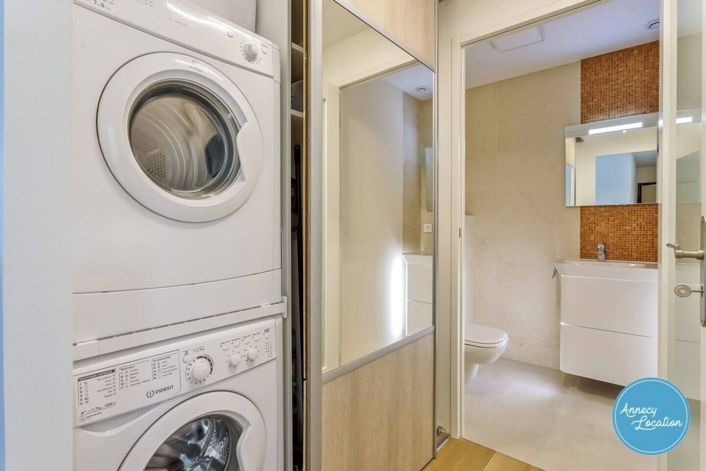 Un espace buanderie avec des machines à laver superposées, une porte vitrée donnant accès à une salle de bain équipée de toilettes et d’un lavabo.