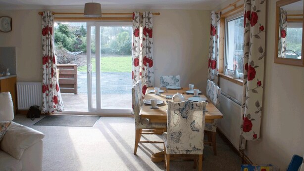 A dining area with a table set for four, surrounded by floral curtains, a wooden bench, and a view of the outdoors. - 3 Bed Bungalow - Sleeps 6 - Pet Friendly - Parking (Stranraer)