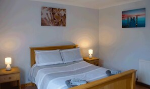 A bedroom with a wooden bed, white bedding, two bedside lamps, and artwork on the walls. - 3 Bed Bungalow - Sleeps 6 - Pet Friendly - Parking (Stranraer)