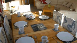 A dining area with a wooden table set for four, surrounded by patterned chairs. A television is mounted on the wall to the left, and a sofa with cushions is in the background. - 3 Bed Bungalow - Sleeps 6 - Pet Friendly - Parking (Stranraer)