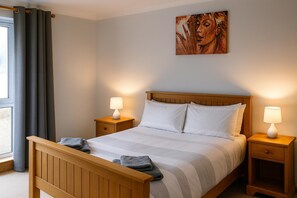 A neatly made bed with a wooden headboard, two bedside tables with lamps, a painting on the wall, and a window with curtains. - 3 Bed Bungalow - Sleeps 6 - Pet Friendly - Parking (Stranraer)