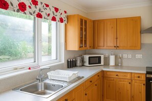 A kitchen with wooden cabinets, a stainless steel sink, a microwave, and a window with a floral curtain. - 3 Bed Bungalow - Sleeps 6 - Pet Friendly - Parking (Stranraer)