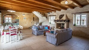 Ein geräumiges Wohnzimmer mit Kamin, gestreiften Sofas, einem Essbereich mit weißen Stühlen und einer Holzbalkendecke.