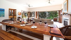 A spacious dining area with a wooden table set for a meal, large windows offering a view of the outdoors, and a cozy seating area with a sofa and chairs. - Tula - waterfront reserve (Currarong)