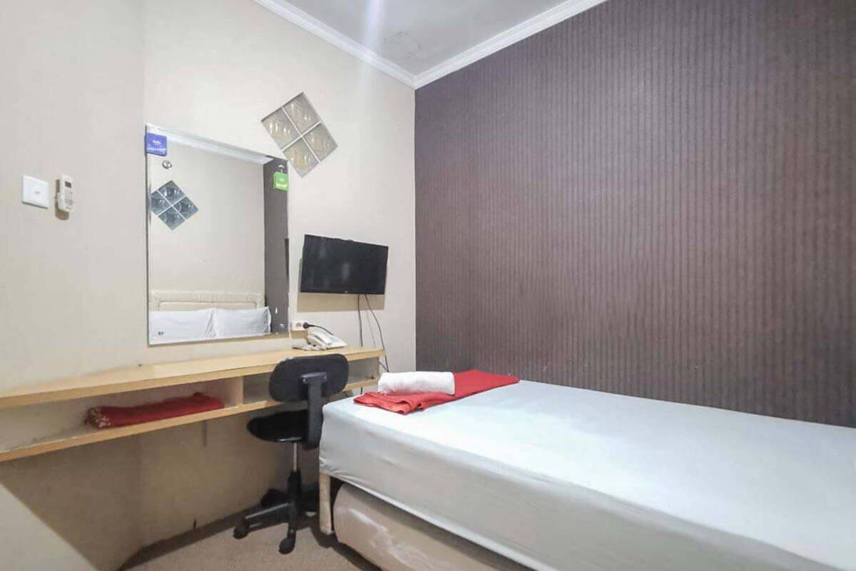 A small, neatly arranged hotel room with a single bed, a desk with a chair, a flat-screen TV, and a small shelf.
