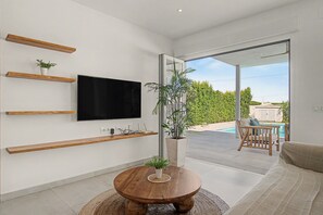 A modern living room with a flat-screen TV, wooden shelves, a round wooden coffee table, and a view of a pool and outdoor seating. - Casa Robert in Conil with Pool, Hot Tub, Garden, and Wi-Fi (Conil)