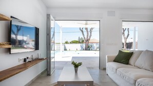 A modern living room with a flat-screen TV, a white sofa, a coffee table, and a view of a swimming pool and outdoor area. - Casa Robert in Conil with Pool, Hot Tub, Garden, and Wi-Fi (Conil)