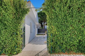 Un étroit sentier bordé de haies verdoyantes et touffues, qui mène à un bâtiment blanc doté d’une porte bien visible et d’une petite fenêtre.