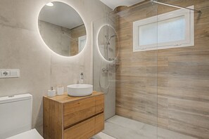 Une salle de bain moderne avec un miroir rond, un meuble-lavabo en bois, un lavabo blanc et une douche séparée par une paroi de verre.