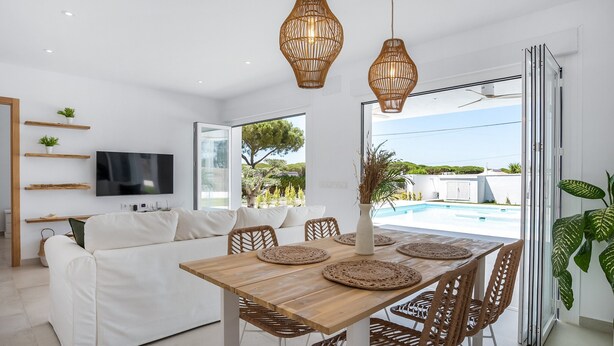 A modern living room with a white sofa, wooden dining table, wicker chairs, and a TV mounted on the wall. - Casa Robert in Conil with Pool, Hot Tub, Garden, and Wi-Fi (Conil)