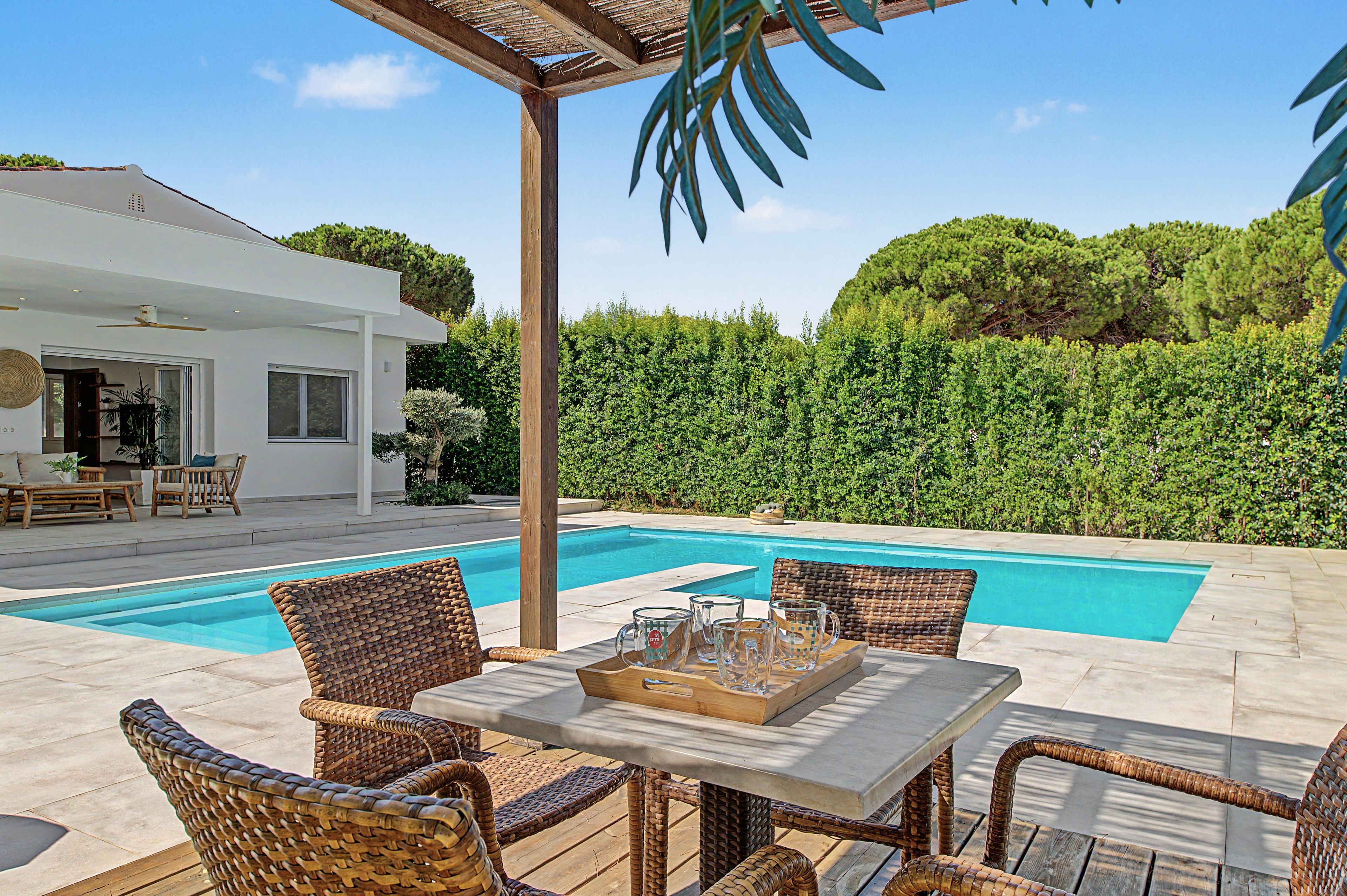 Área de piscina com mobiliário de vime e uma mesa posta para dois, cercada por um viveiro e uma casa ao fundo.