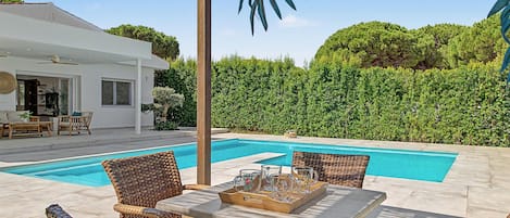 Zona junto a la piscina con mobiliario de mimbre y una mesa dispuesta para dos personas, rodeada por un seto y una casa al fondo.