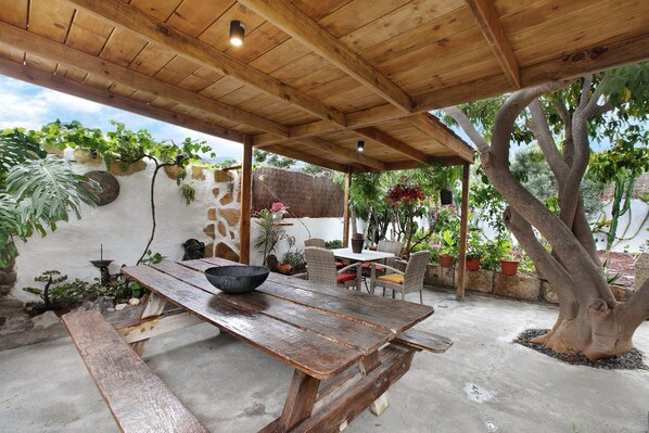 A wooden pergola with a table and chairs, surrounded by potted plants and a tree.