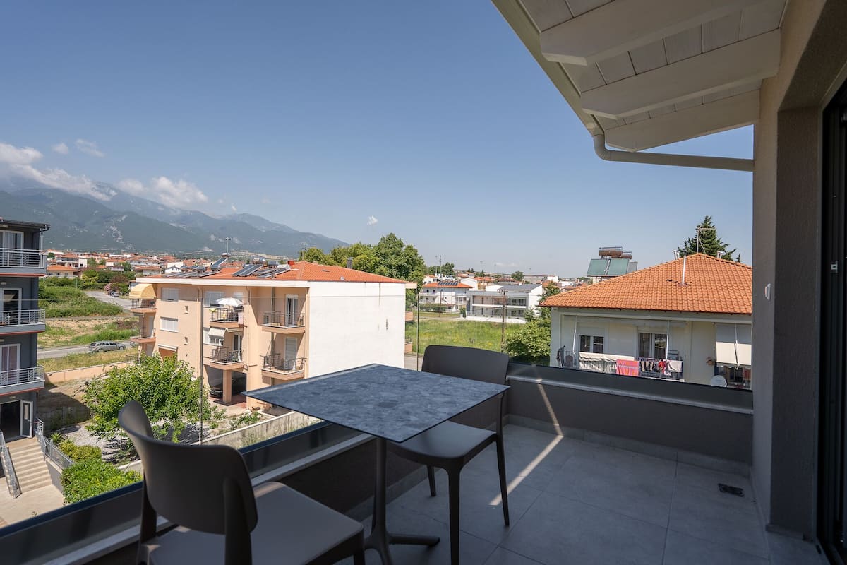 A balcony with a table and chairs, overlooking a residential area with houses and mountains in the background.