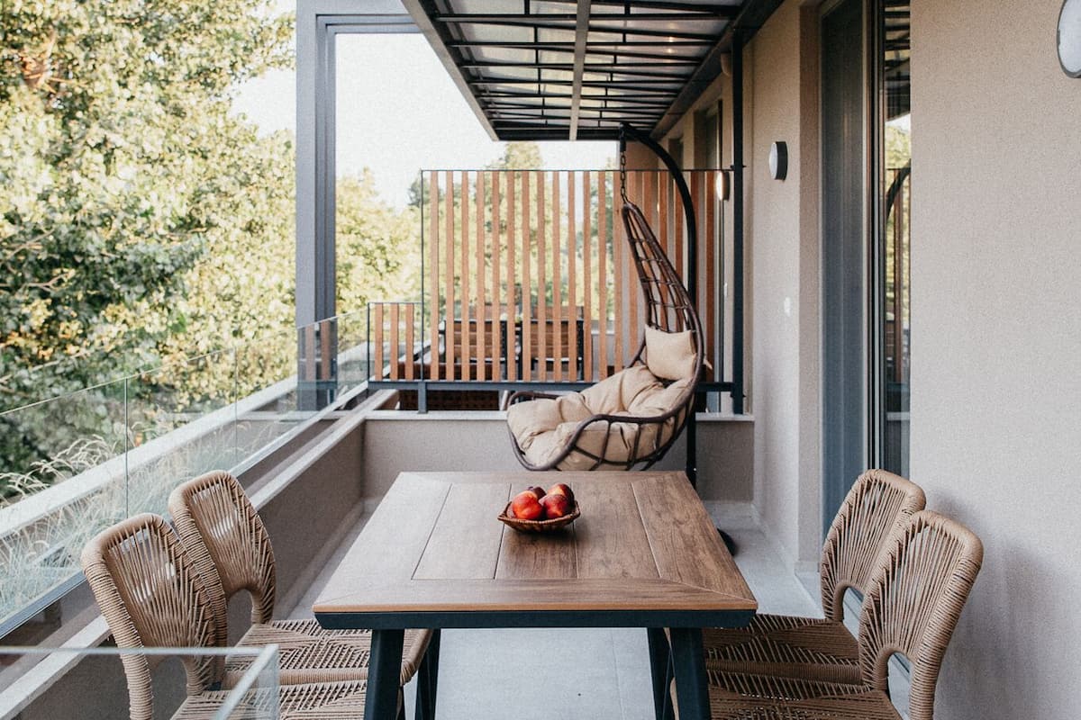 A modern outdoor dining area with a wooden table and wicker chairs, a hanging chair, and a glass railing overlooking greenery.