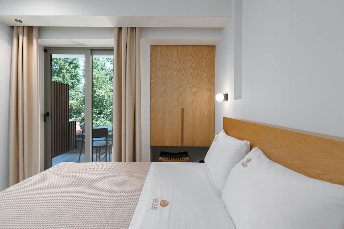 A hotel room with a large bed, a wooden headboard, a view of the outdoors, and a sliding door leading to a balcony.