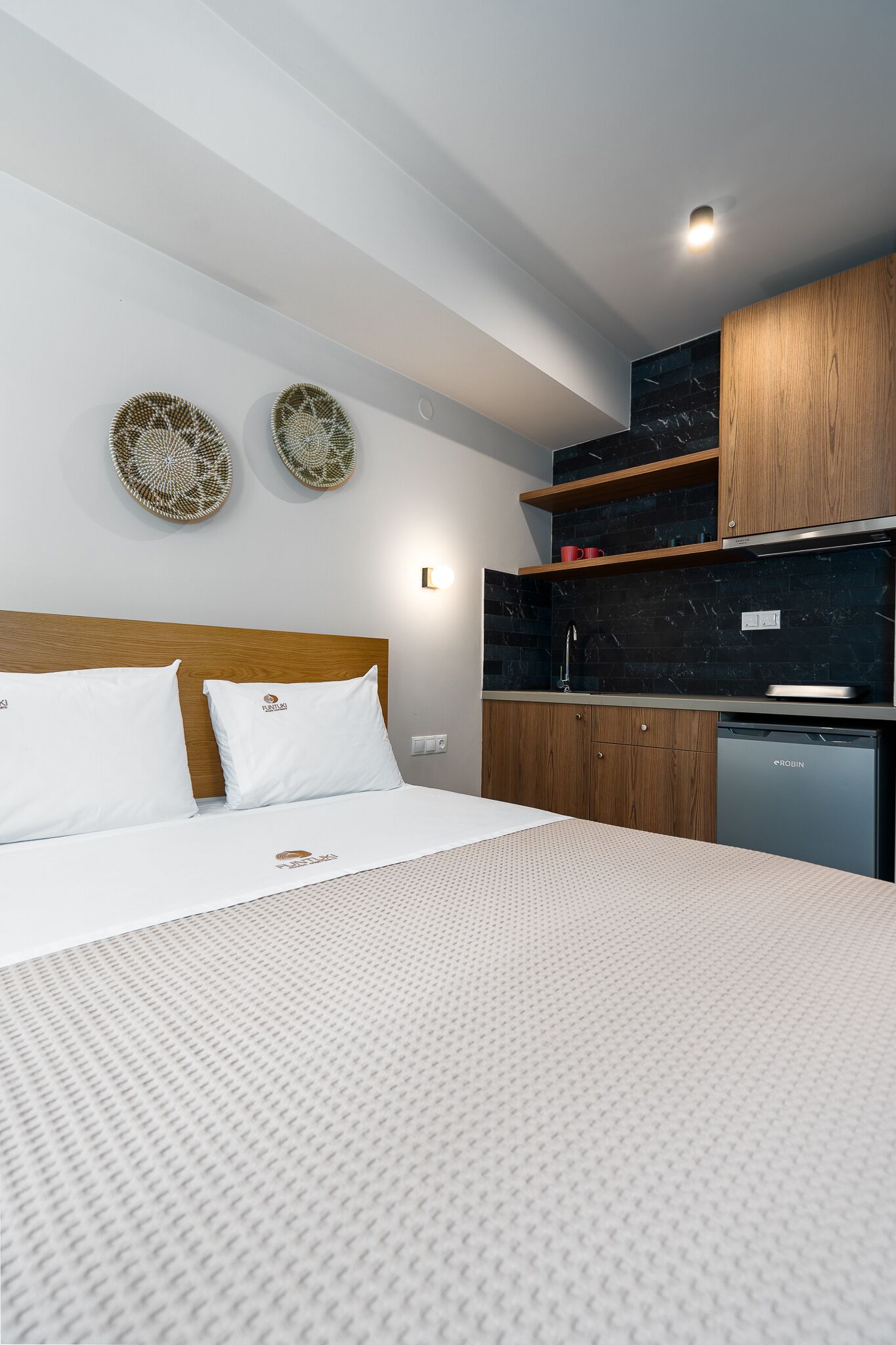 Une chambre d’hôtel moderne avec un lit soigneusement fait, une tête de lit en bois, une cuisine intégrée équipée d’un lave-vaisselle et des œuvres d’art décoratives au mur.