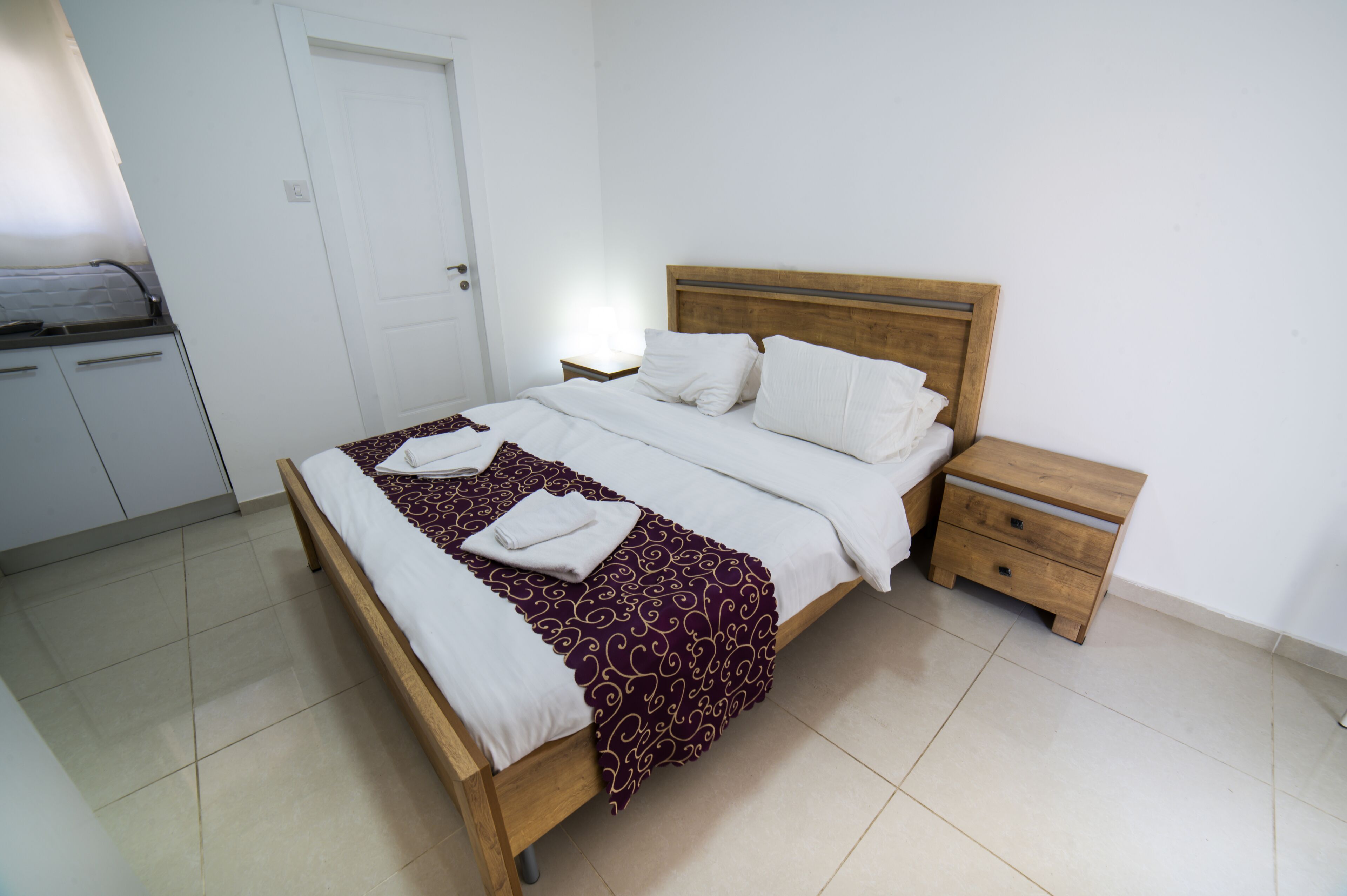 Un lit bien fait, avec une tête de lit en bois, une table de chevet et une porte blanche, dans une pièce propre et au décor minimaliste.