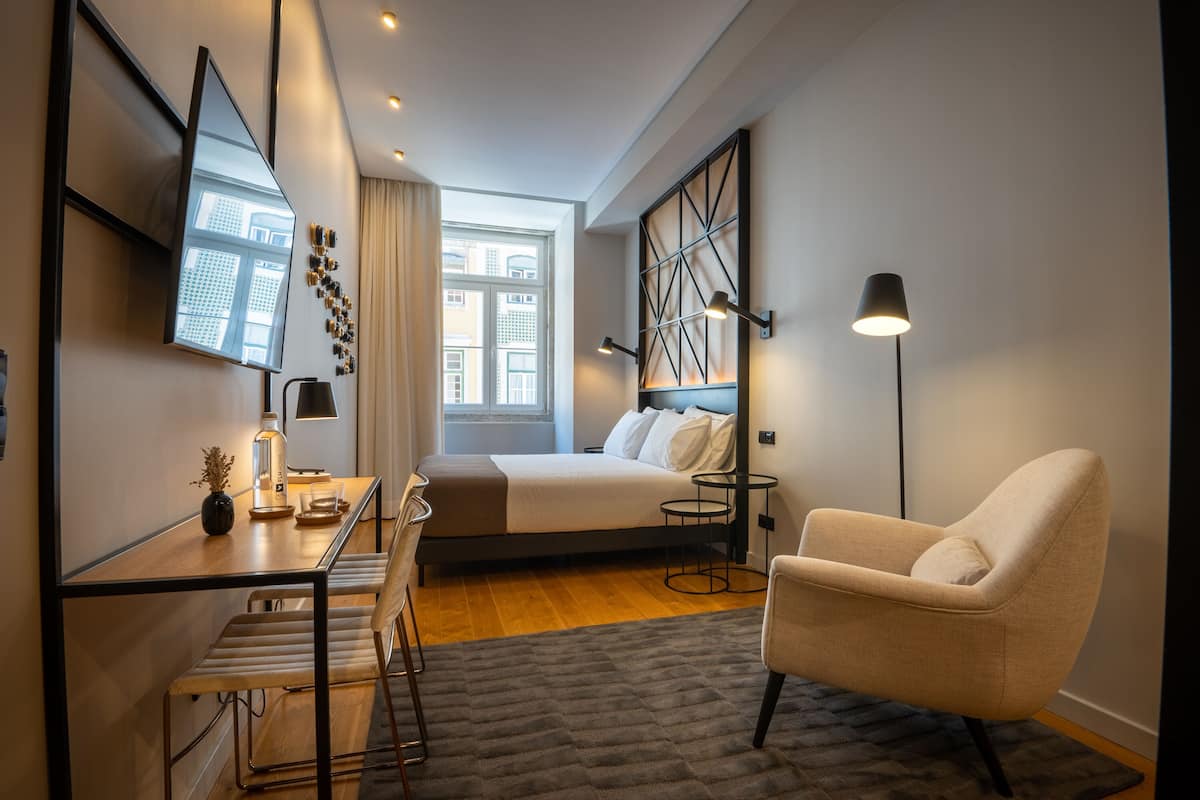 Una habitación de hotel moderna con cama, escritorio, silla, lámpara y un amplio ventanal con vistas a edificios.