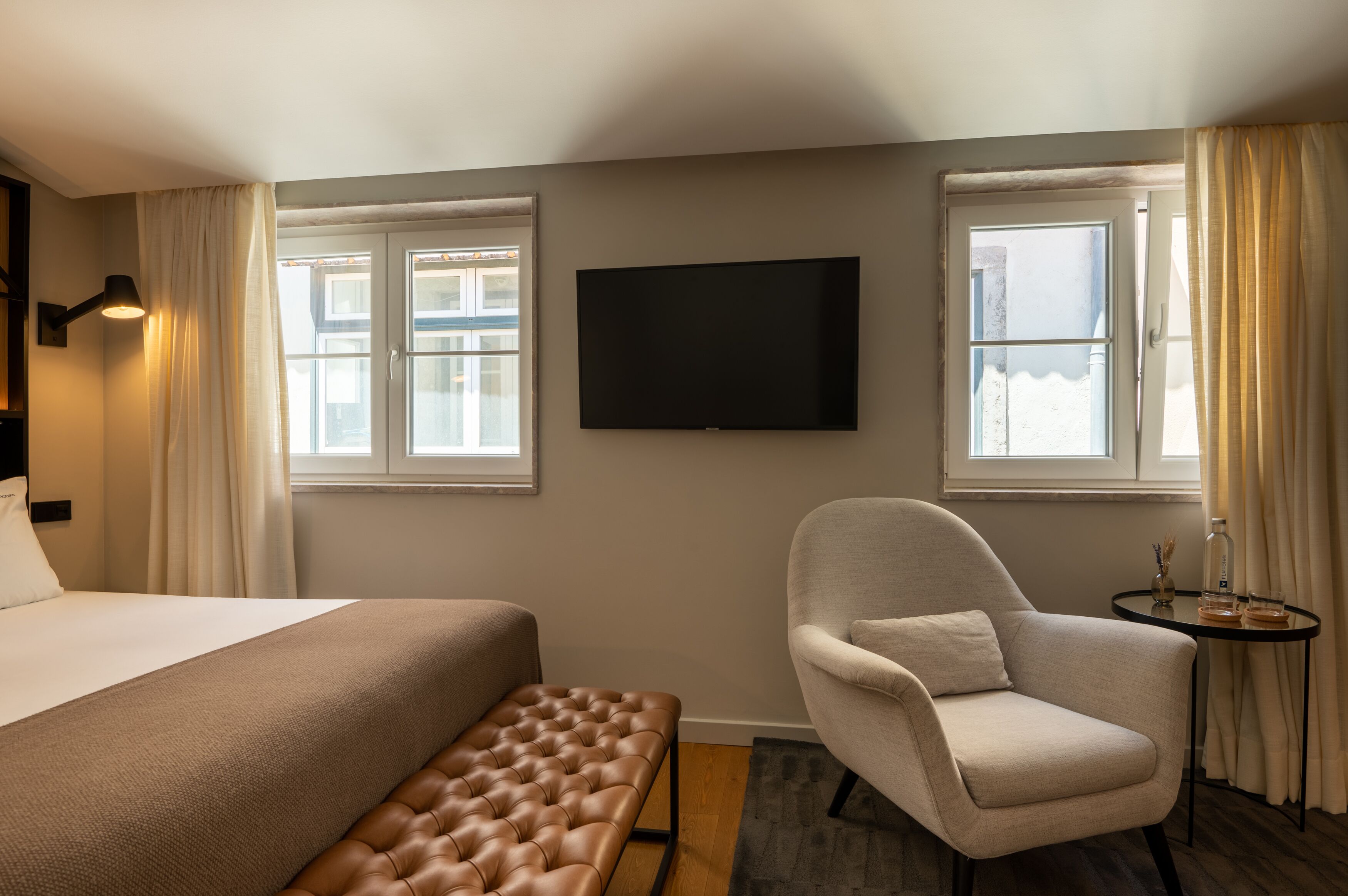A hotel room with a bed, a chair, a small table with a bottle, and a TV on the wall.