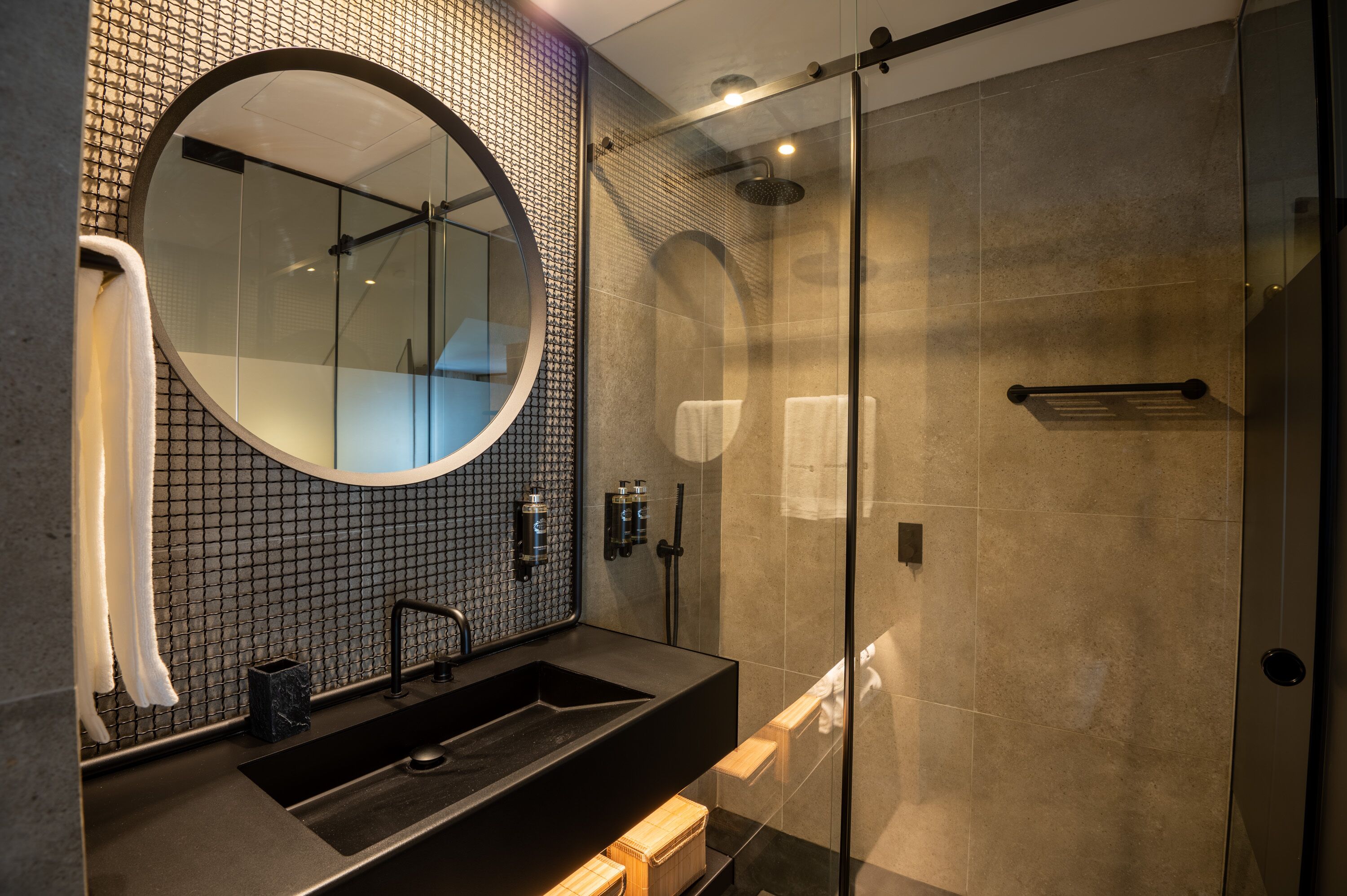 Un baño moderno con un espejo redondo grande, un lavamanos negro y una zona de ducha con mampara de cristal.