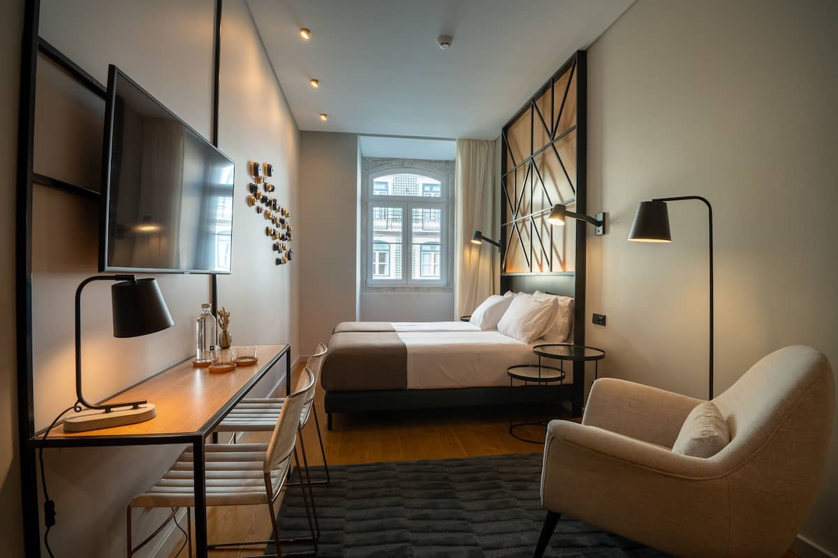Habitación de hotel moderna con cama, escritorio, silla y un ventanal con vistas a edificios.