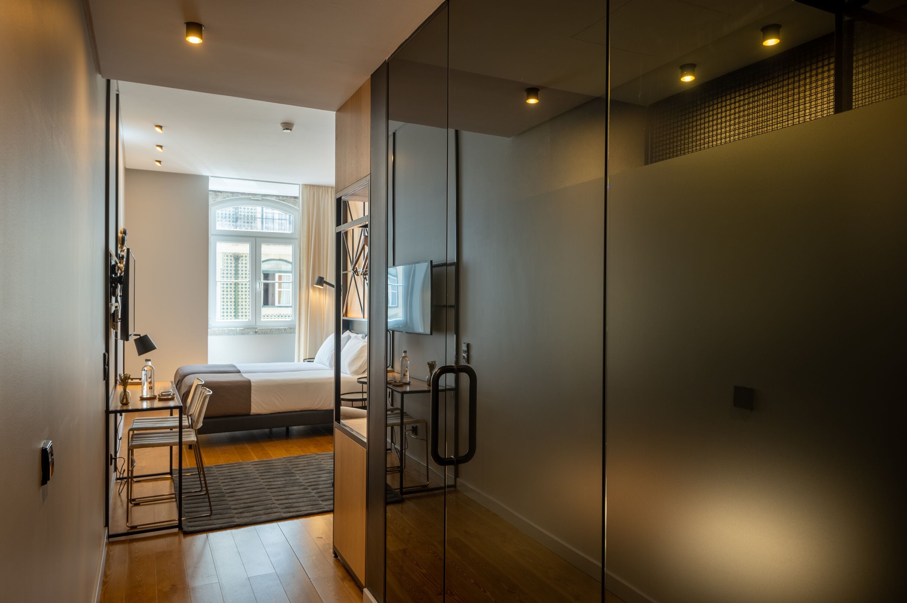 A modern hotel room with a bed, bedside table, lamp, and a view of the city through a window.