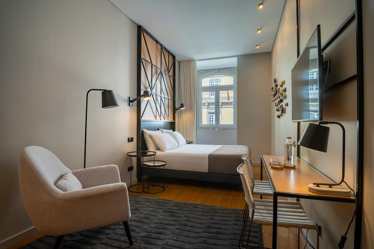 Una habitación de hotel moderna con una cama, un escritorio, una silla, una lámpara y vistas a edificios por la ventana.