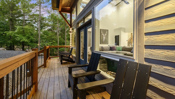Eine Holzterrasse mit Adirondack-Stühlen, eine Glastür, die zum Innenbereich führt, und Blick auf den umliegenden Wald.