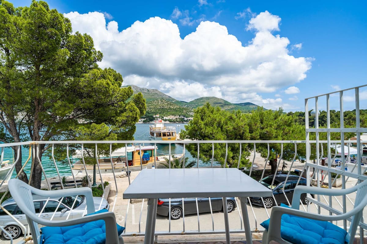 Terraço com mesa e cadeiras, com vista para uma marina com barcos e montanhas ao fundo.