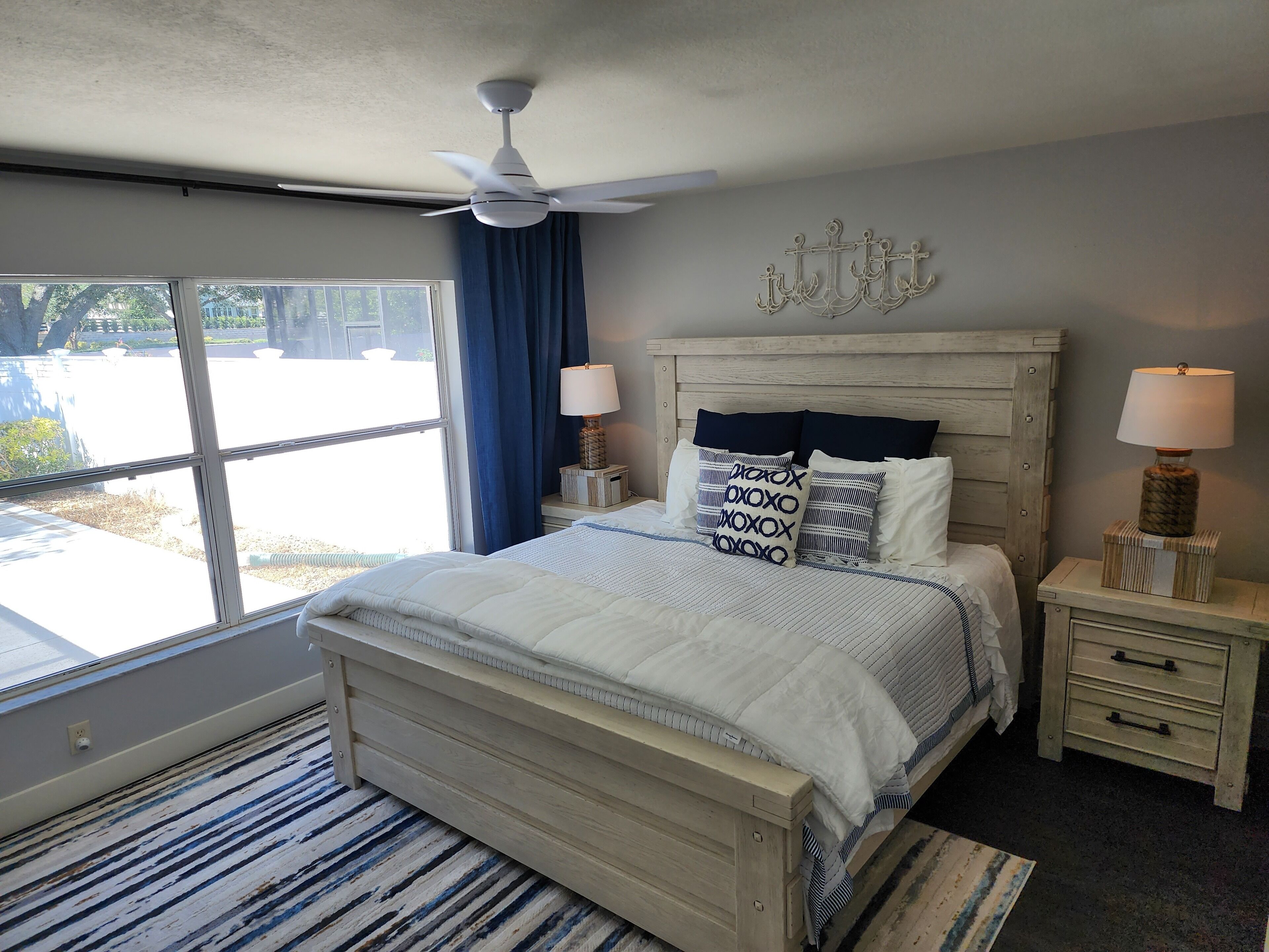 A bedroom with a bed, two bedside tables, a ceiling fan, a window with curtains, and a view of the outside.