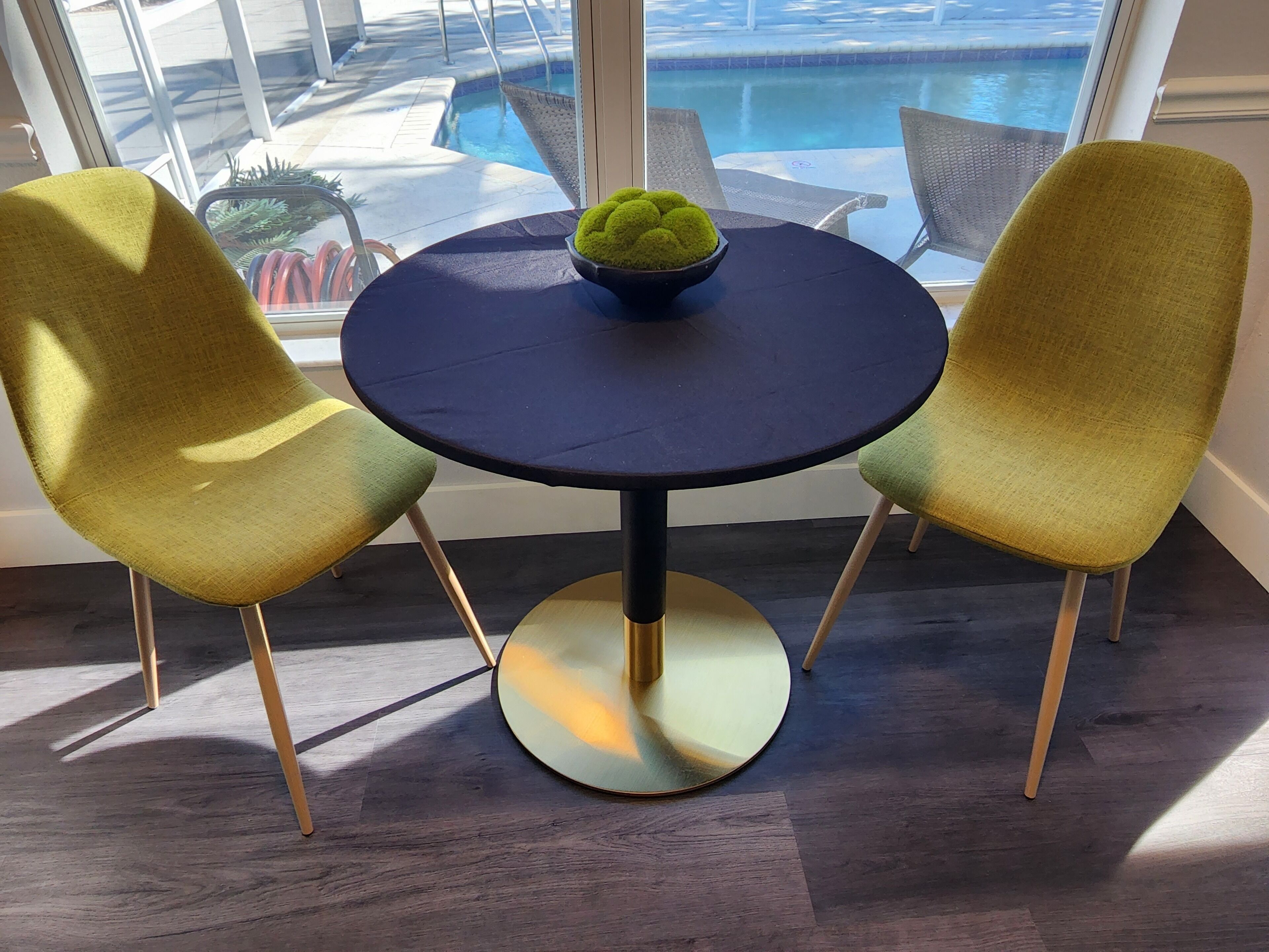 A round table with a dark tablecloth, two green chairs with gold legs, and a bowl of green spherical objects on the table.