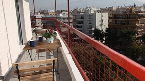 Un balcon avec une table en bois, deux plantes en pot et une rambarde rouge donnant sur un paysage urbain avec des bâtiments et des montagnes à l’horizon.