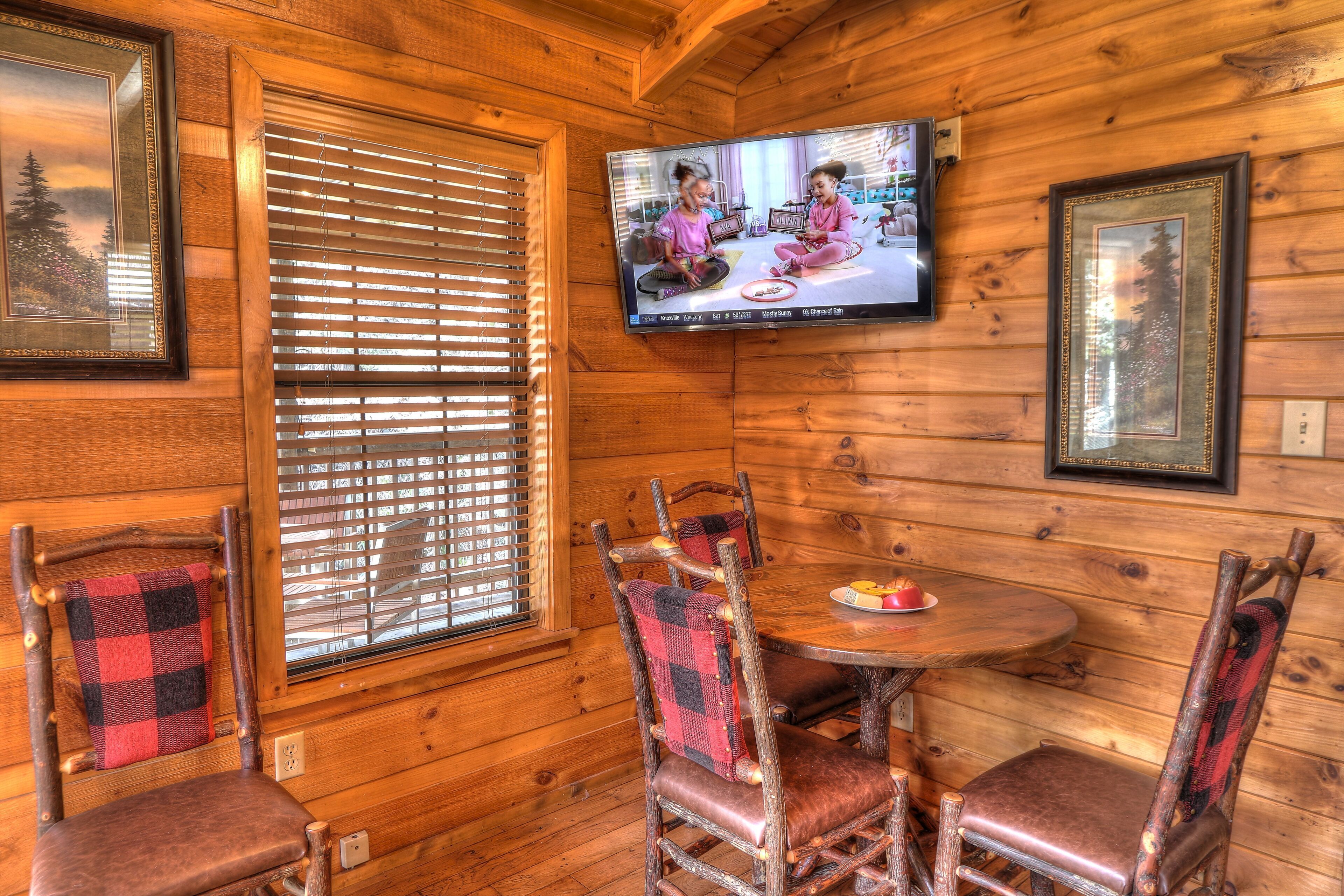A cozy wooden cabin interior with a television mounted on the wall, a dining table with chairs, and framed pictures on the walls.