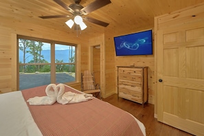 Ein Zimmer in einer Holzhütte mit einem Bett, einem Deckenventilator, einem Fernseher und Blick ins Freie.