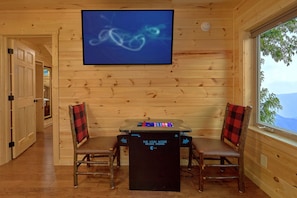 Ein Holzblockhaus-Innenraum mit einem an der Wand befestigten Fernseher, der ein Spiel zeigt, einem Tisch mit einer Spielkonsole, zwei karierten Sesseln und einem Fenster mit Blick.