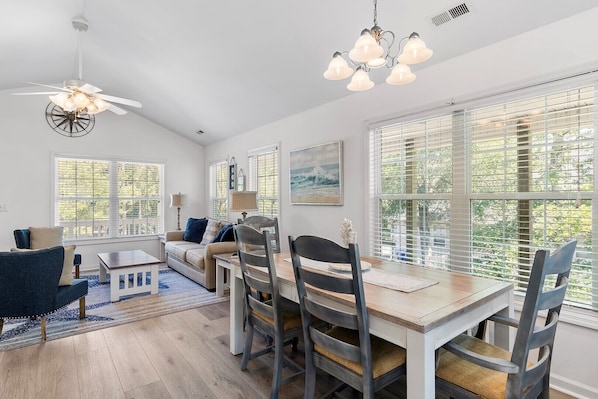 A spacious living room with a dining area, large windows with blinds, a ceiling fan, and a chandelier.