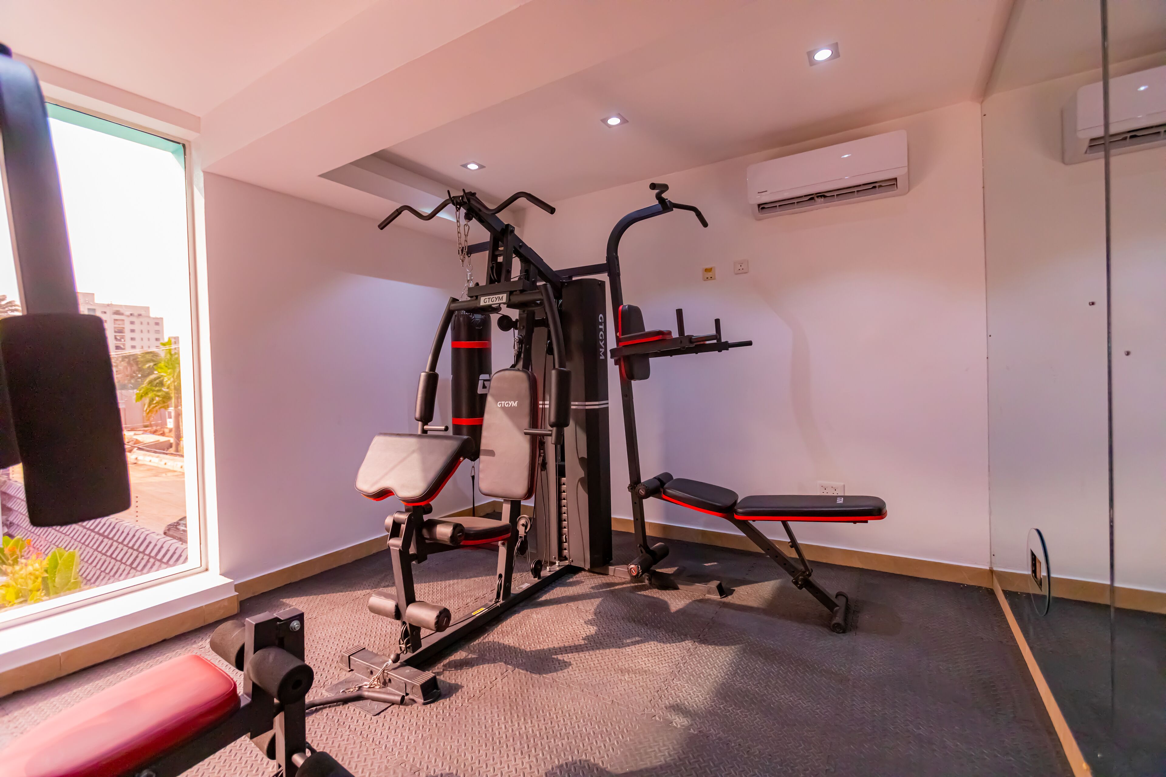 A gym with exercise equipment, a bench, and a window with a view of buildings and trees.