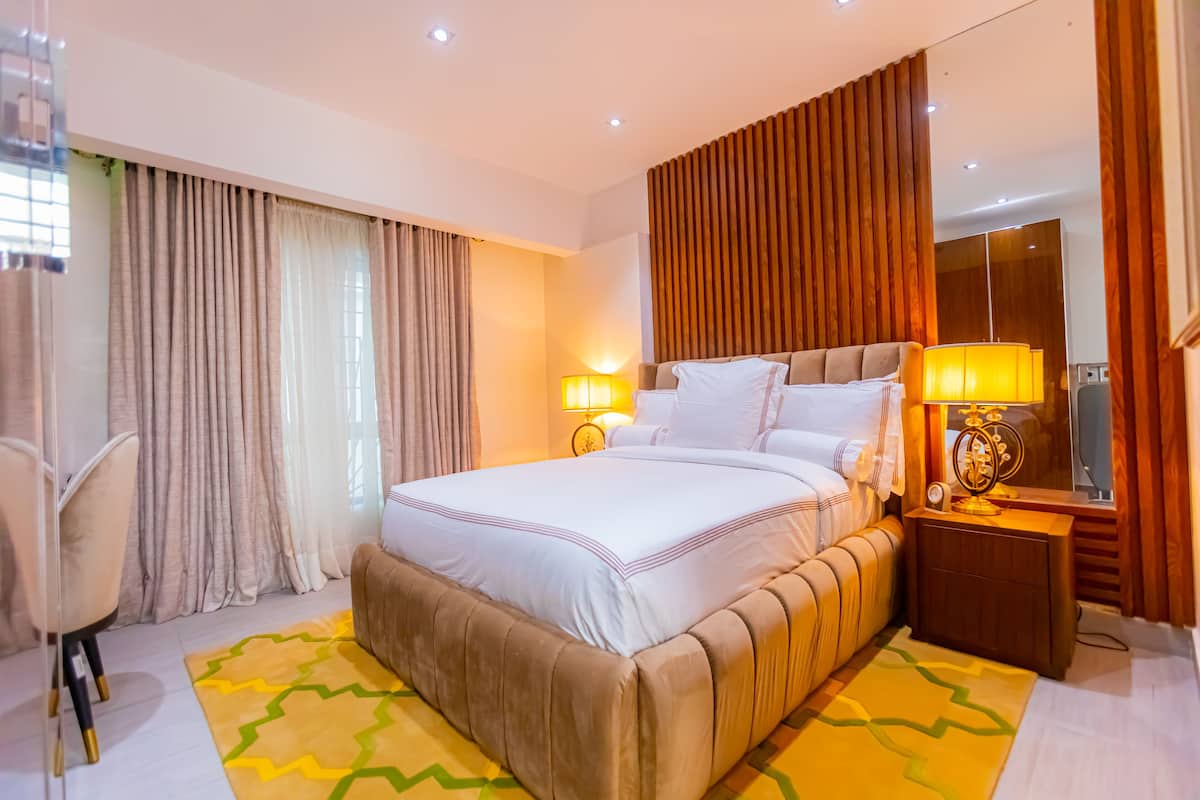 A modern hotel room with a large bed, a wooden headboard, a nightstand with a lamp, and a large mirror.
