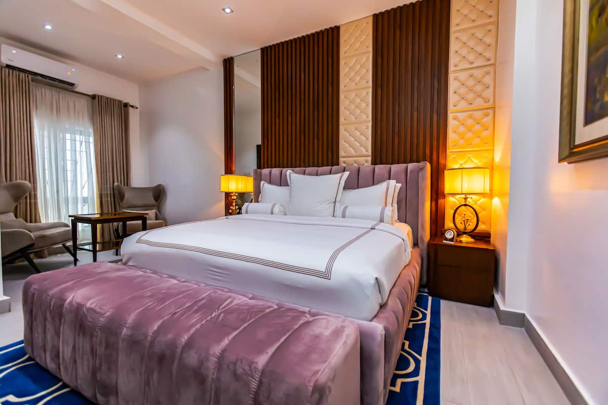 A hotel room with a large bed, a sitting area with a chair and a small table, and a bedside table with a lamp.