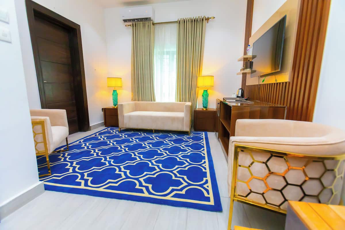 A hotel room with a blue patterned carpet, a sofa, a chair, a TV, and a bathroom with a white sink and mirror.