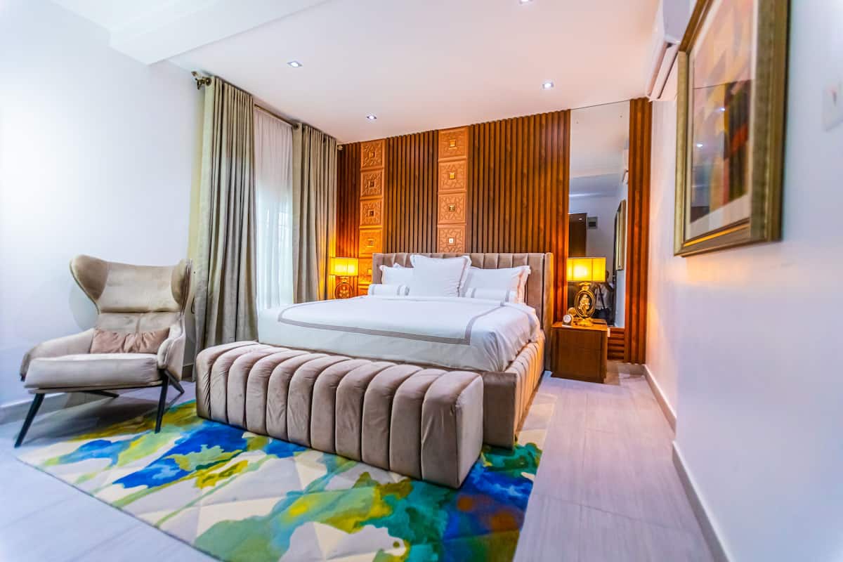 A modern hotel room with a large bed, a chair, a bedside table, and a colorful rug.