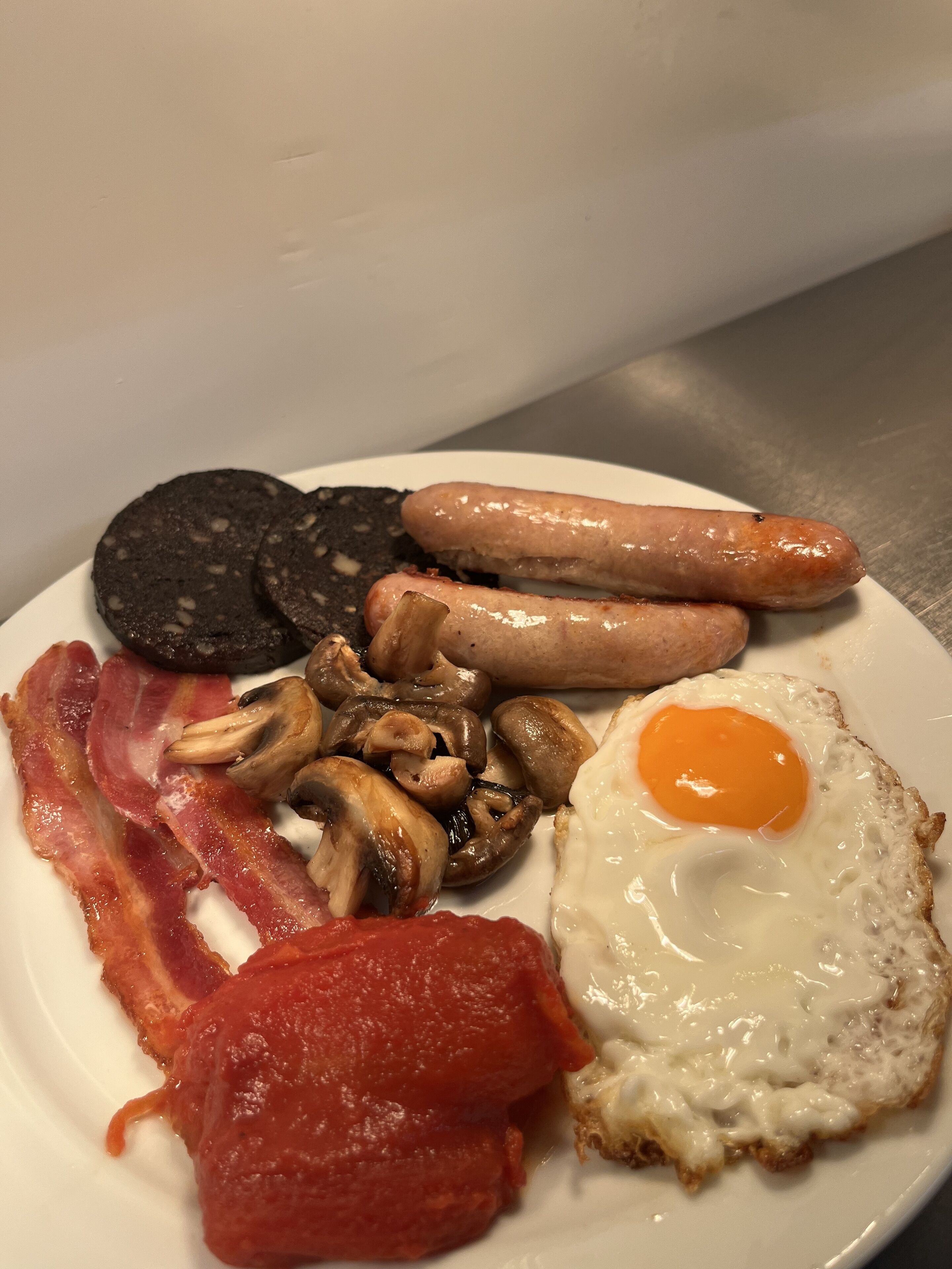 A plate of breakfast food including sausages, mushrooms, bacon, a fried egg, and tomato sauce.