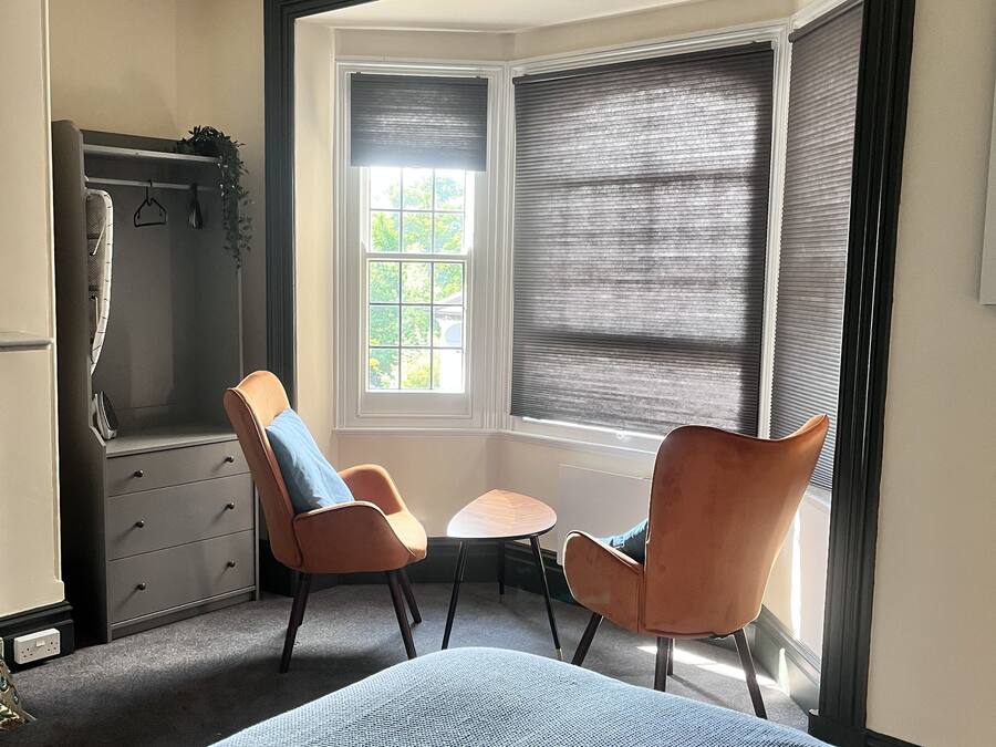 A bedroom with a large bed, two chairs, a small table, a wardrobe, and a window with blinds.