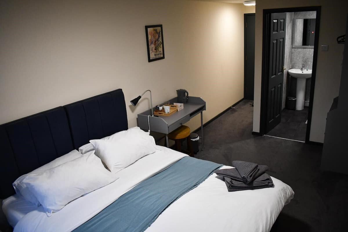 A hotel room with a bed, a desk, a chair, a small table with a cup and a kettle, and a bathroom visible through an open door.
