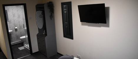 Una habitación de hotel con cama, televisor montado en la pared, baño con inodoro y lavabo, y puerta que da a otra habitación.