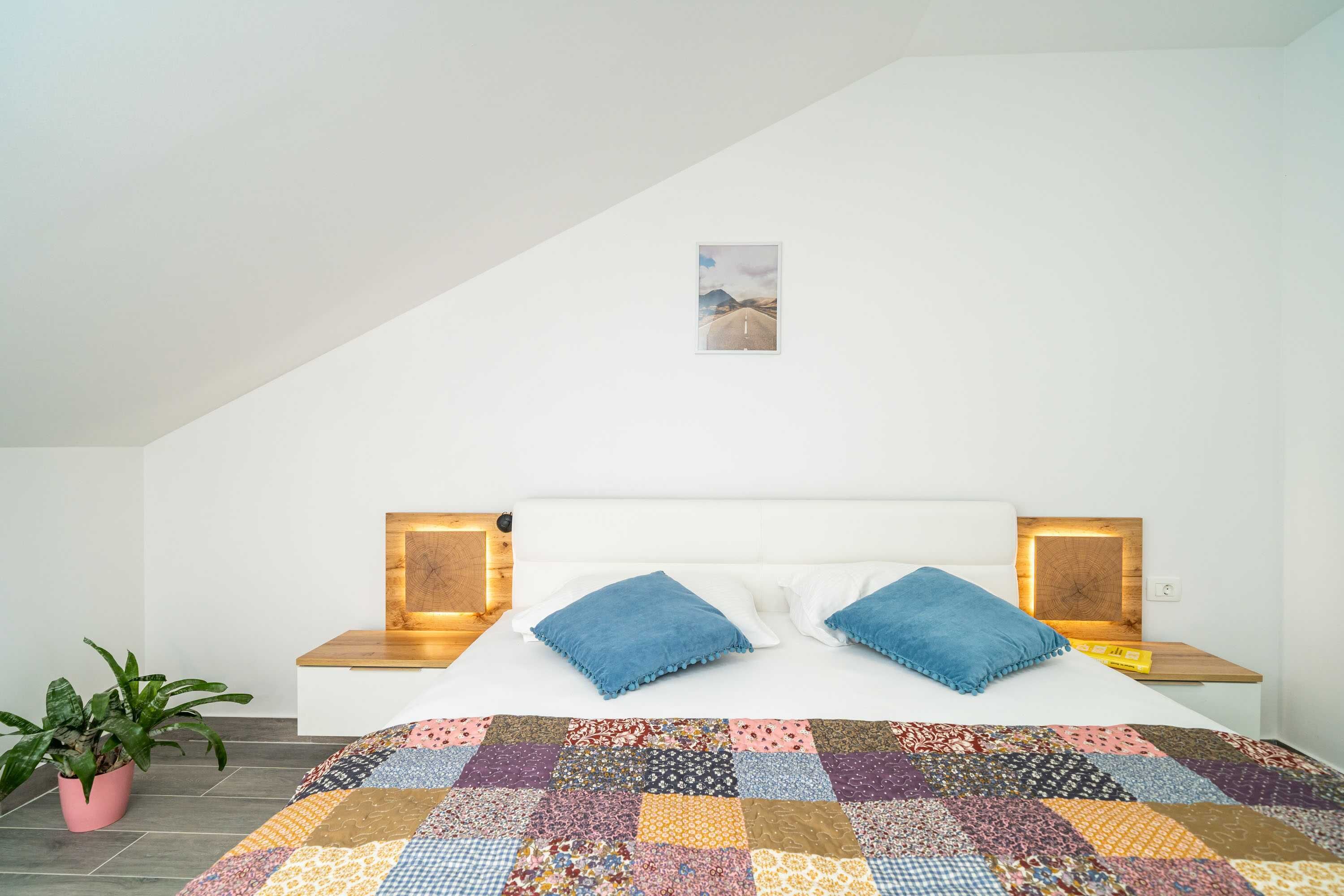 Quarto com cama grande, cabeceira de madeira, duas mesas de cabeceira com abajures, um vaso de planta e um pequeno quadro na parede.