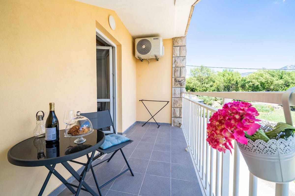 A balcony with a table, a chair, a bottle of wine, a glass, a basket with flowers, and an air conditioning unit.